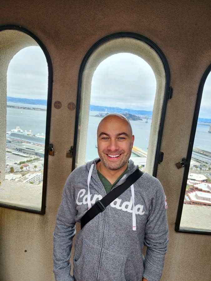 Manny smiling in a Canada hoodie inside a tower with arched windows overlooking a bay
