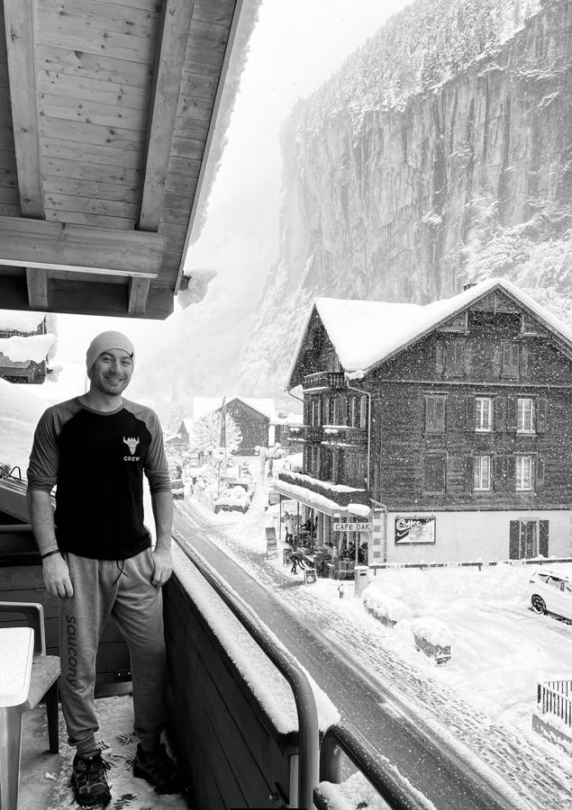 Black-and-white photo of Manny on a snowy chalet balcony with a cliff behind him