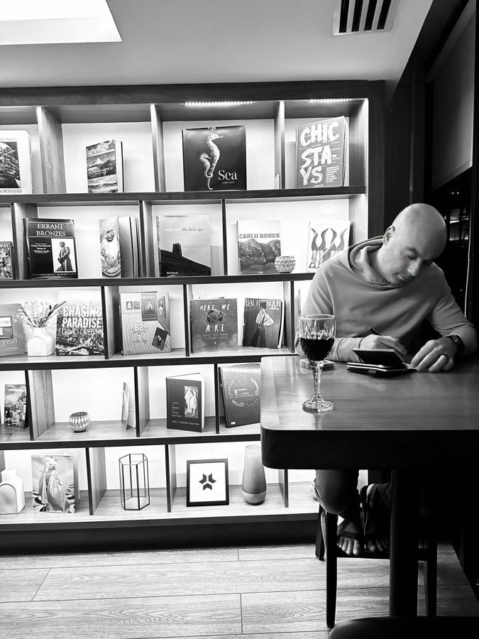 Black-and-white photo of Manny writing in a notebook beside a glass of red wine in front of a lit bookcase